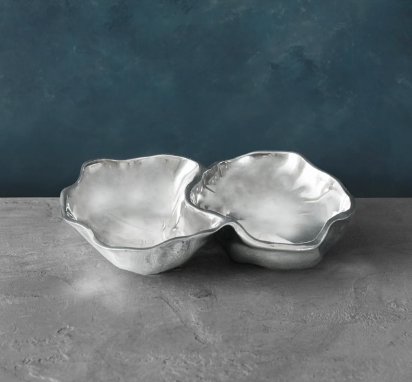 A shiny, silver Beatriz Ball Soho Onyx Double Dip Serving Bowl with wavy edges rests on a gray textured surface against a blue marbled background—ideal for serving dips or small bites.