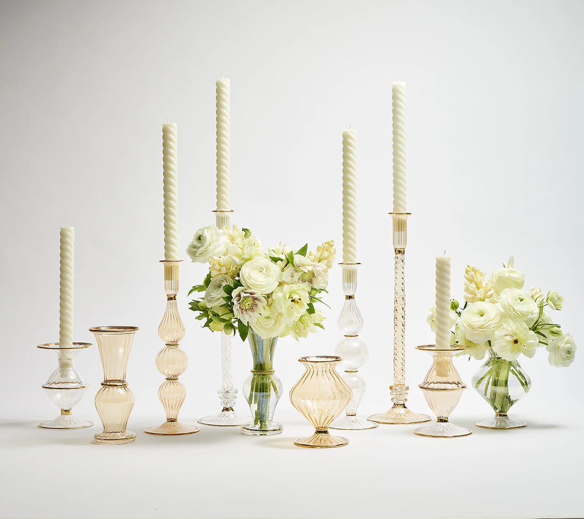 A set of Kim Seybert Champagne Rib Gold Detail Vases filled with white flowers, paired with elegant glass candlesticks and tall white candles, arranged on a white surface against a plain white background.