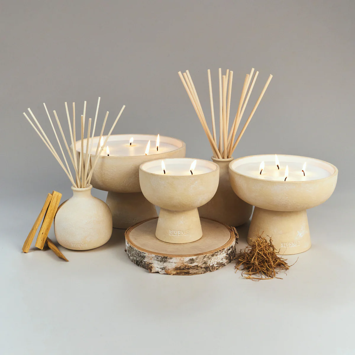 Blueme’s Spiritual Candle, crafted from a soy wax blend in a beige ceramic vessel, is styled with reed diffusers on light wood slices and dried botanicals, set against a neutral gray background for subtle wellness fragrances.
