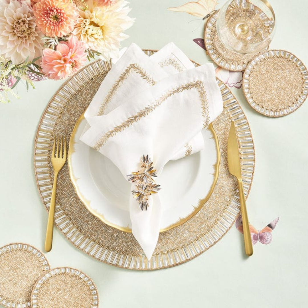 Elegant table setting with gold utensils, a white plate with gold trim, embroidered napkins accented by Kim Seybert’s Butterflies Napkin Ring Set of 4, gold and beaded placemats and coasters, a glass of wine, and pink and cream flowers.