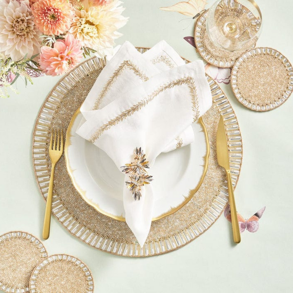 Elegant table setting with gold utensils, a white plate with gold trim, embroidered napkins accented by Kim Seybert’s Butterflies Napkin Ring Set of 4, gold and beaded placemats and coasters, a glass of wine, and pink and cream flowers.