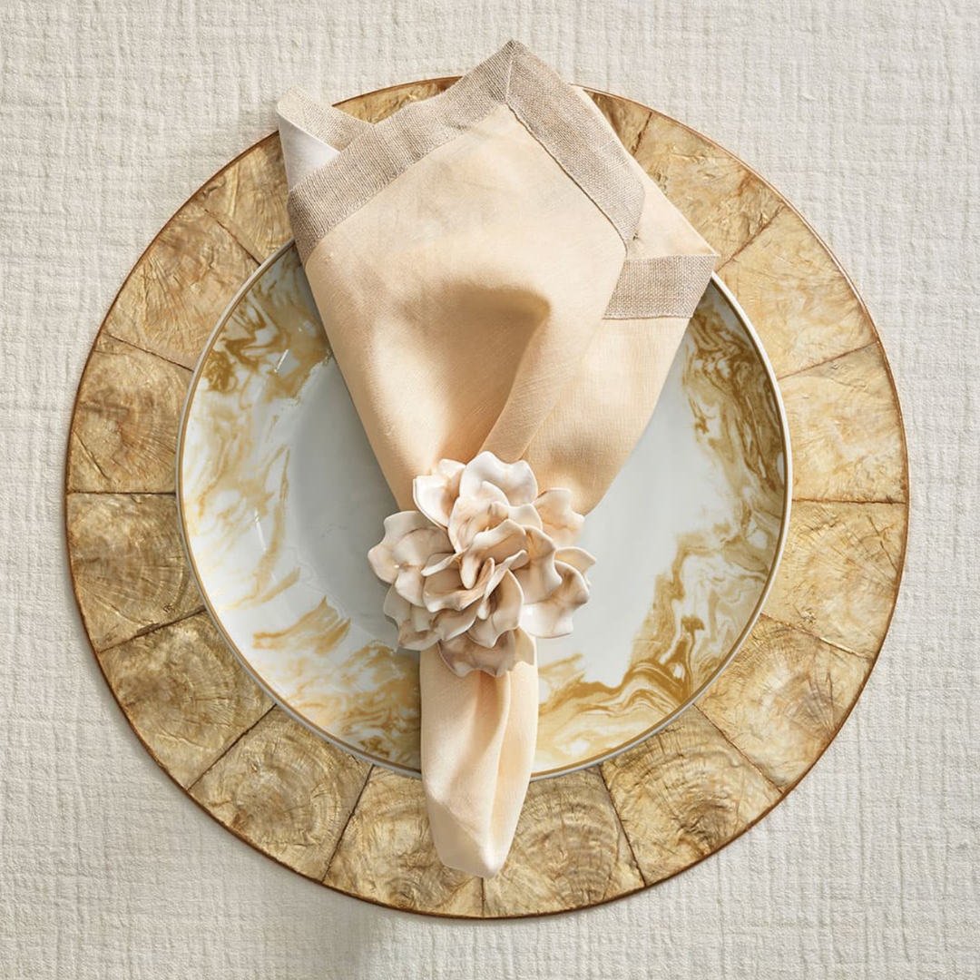 A beige napkin with a floral ring sits on a marbled plate with gold accents atop a textured gold charger, all layered on the Kim Seybert Capiz Shell Placemat Champagne Set of 4, over a light fabric tablecloth.