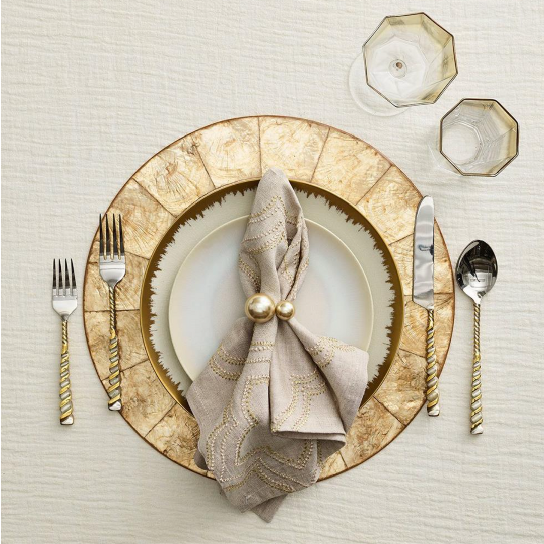 An elegant place setting features a Kim Seybert Capiz Shell Placemat Champagne, a white plate, tan napkin with gold embroidery held by two gold balls, and silverware with gold twisted handles.