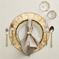An elegant place setting features a Kim Seybert Capiz Shell Placemat Champagne, a white plate, tan napkin with gold embroidery held by two gold balls, and silverware with gold twisted handles.