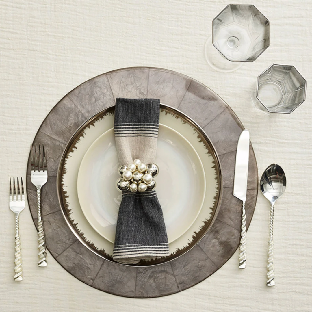 A formal table is set with a plate on the Kim Seybert Capiz Shell Placemat Grey Set of 4, a napkin with a pearl ring, twisted silver cutlery, and two faceted glasses on a light textured tablecloth.
