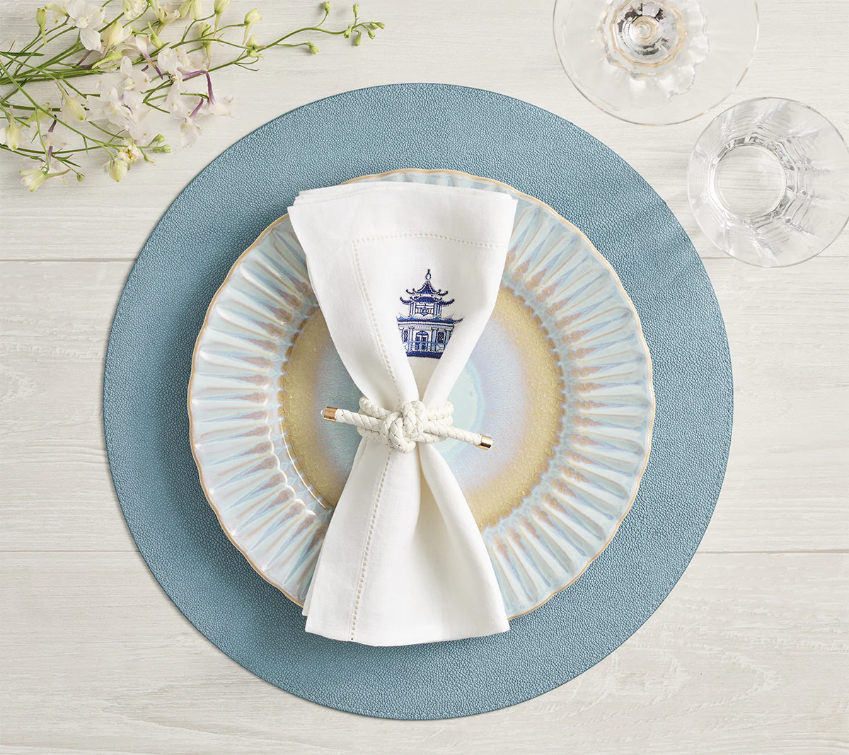 A Kim Seybert Icons Placemat from the Set of 4 in light blue shagreen texture is on a white wood table, topped with a decorative plate, knotted white napkin, floral glass, wine glass, and small flowers.