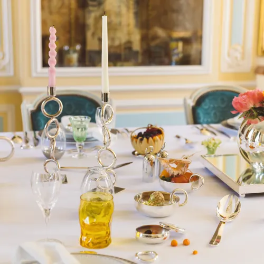 Elegant table setting featuring Christofle Vertigo Silver Plated Candlesticks with pink and white candles, a yellow drink, assorted desserts, glassware, and pink flowers in a vase on a white tablecloth in a luxurious room.