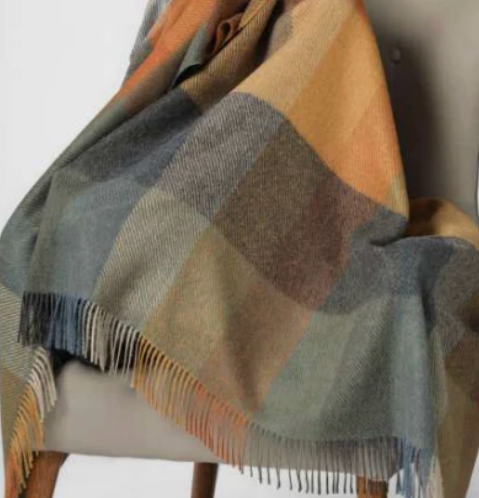 The Sloane Alpaca Throw by Alicia Peru Home, featuring color blocks in gray, blue, brown, and orange with fringed edges, is draped over the back and seat of a light-colored upholstered chair.