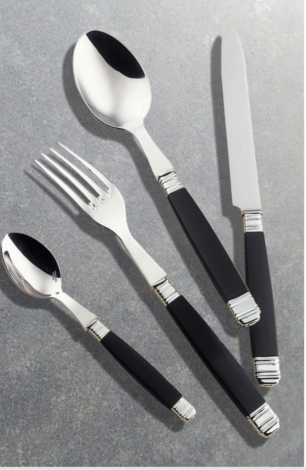 The Alain Saint-Joanis Empire Black Flatware 5 Piece Setting features stainless steel heads and sleek black handles, including a spoon, fork, knife, and teaspoon, stylishly displayed on a gray stone surface.