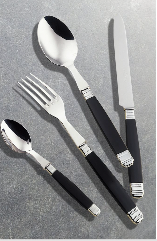 The Alain Saint-Joanis Empire Black Flatware 5 Piece Setting features stainless steel heads and sleek black handles, including a spoon, fork, knife, and teaspoon, stylishly displayed on a gray stone surface.