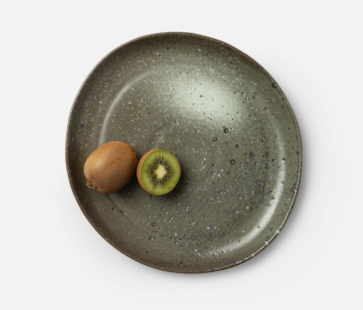 A whole kiwi and a sliced half kiwi sit on the Marcus Gray Salt Dinnerware by Blue Pheasant, a speckled green ceramic plate, against a white background.