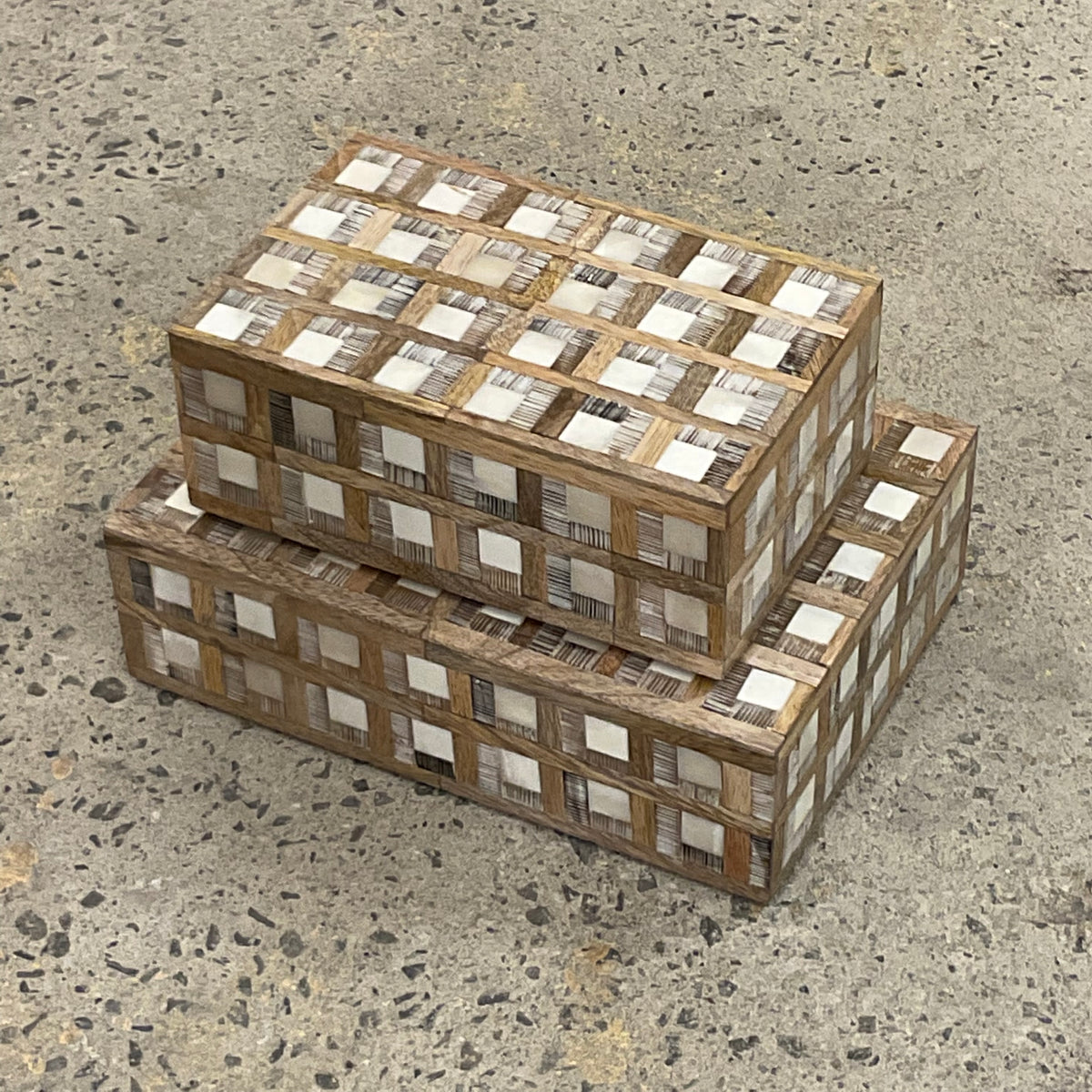 A set of two rectangular Plaid Wood and Bone Boxes by Bojay—featuring distinctive checkered patterns in contrasting bone inlay and rustic wood—are stacked atop a speckled concrete floor, lending a refined, masculine touch to the space.