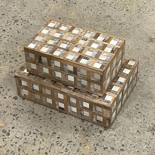 A set of two rectangular Plaid Wood and Bone Boxes by Bojay—featuring distinctive checkered patterns in contrasting bone inlay and rustic wood—are stacked atop a speckled concrete floor, lending a refined, masculine touch to the space.