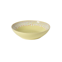 The Costa Nova Taormina Dinnerware Yellow bowl, with its shallow, round shape and wavy orange and blue rim design, showcases contemporary ceramic tableware against a plain white background.