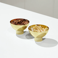 The Anna New York Striations Gold Nut Bowls—two stainless steel bowls with ribbed pedestal bases—are displayed on a white surface, one filled with candied pecans and the other with dried mango slices.