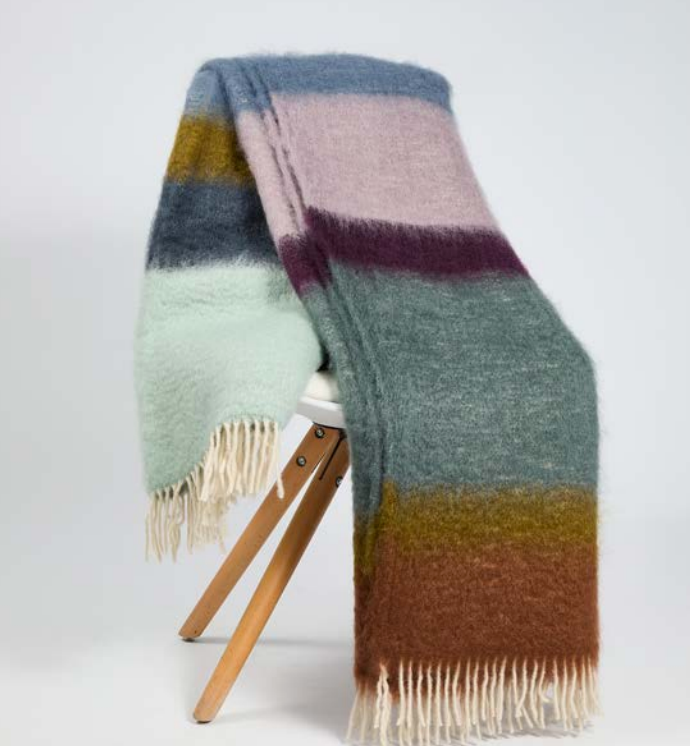 Matisse Teal & Lavender Throw: a fuzzy mohair blanket with multicolored horizontal stripes in teal, lavender, brown, and green, draped over a chair with cream-colored fringe at both ends.