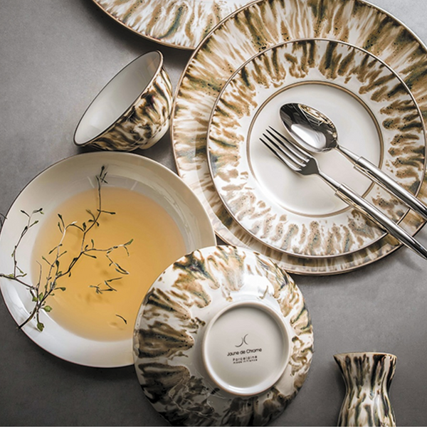 A set of Ecaille Creme Dinnerware by JL Coquet, including patterned porcelain dishes, bowls (one with herb broth), and a small vase, is arranged on a gray surface. Some plates feature silverware.