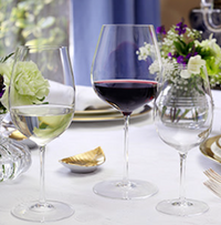 Three William Yeoward Starr Stemware lead-free crystal wine glasses, one with red wine, are arranged on a white tablecloth with elegant white and purple flowers, plates, gold cutlery, and a folded napkin.