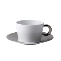 The Bernardaud Ecume Platinum teacup, featuring a white porcelain body and metallic handle, rests on a textured platinum-rimmed saucer—showcasing modern elegance from the Ecume Platinum Dinnerware collection against a white backdrop.