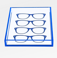 The Eye Candy Specs Eyeglass Tray is a transparent acrylic holder with a blue border, designed to display four pairs of blue eyeglass frames in parallel rows on a white background.