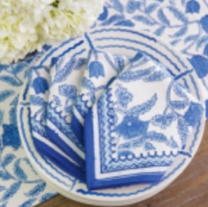 The Pomegranate Jalore Napkin Set of 4 - Blue, made from 100% cotton with a blue floral design, is displayed folded on a white plate over matching table linens and a blue-and-white tablecloth, with white flowers in the corner.