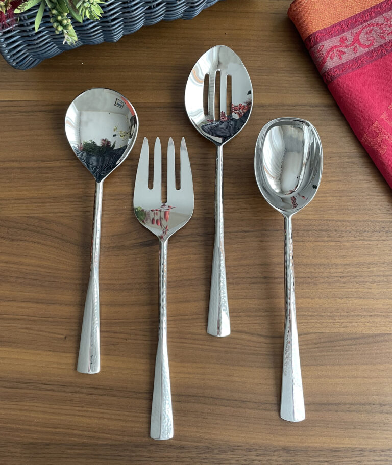 The Mary Jurek Alta Slotted Serving Spoon, part of a four-piece stainless steel serving set, is displayed with other utensils on a wooden table featuring a red cloth and a flower basket in the background.