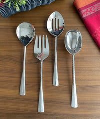 The Mary Jurek Alta Slotted Serving Spoon, part of a four-piece stainless steel serving set, is displayed with other utensils on a wooden table featuring a red cloth and a flower basket in the background.