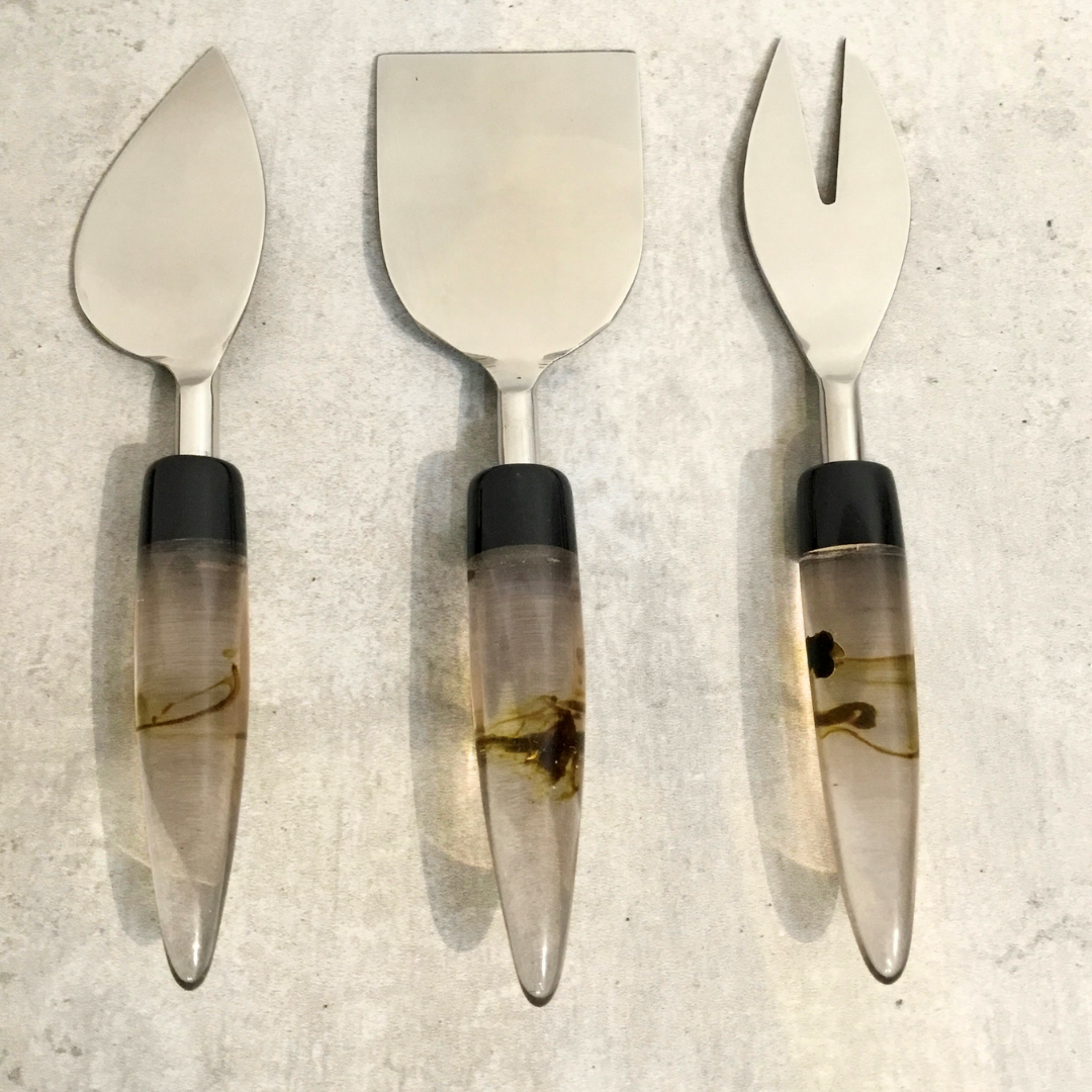 The India Handicrafts Lava Handle Cheese Knives Set features three knives—pointed, rectangular, and forked—with clear lava-inspired handles and brown swirls, arranged vertically on a light gray textured surface.