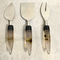 The India Handicrafts Lava Handle Cheese Knives Set features three knives—pointed, rectangular, and forked—with clear lava-inspired handles and brown swirls, arranged vertically on a light gray textured surface.