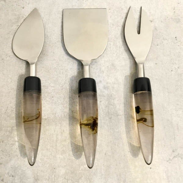 The India Handicrafts Lava Handle Cheese Knives Set features three knives—pointed, rectangular, and forked—with clear lava-inspired handles and brown swirls, arranged vertically on a light gray textured surface.