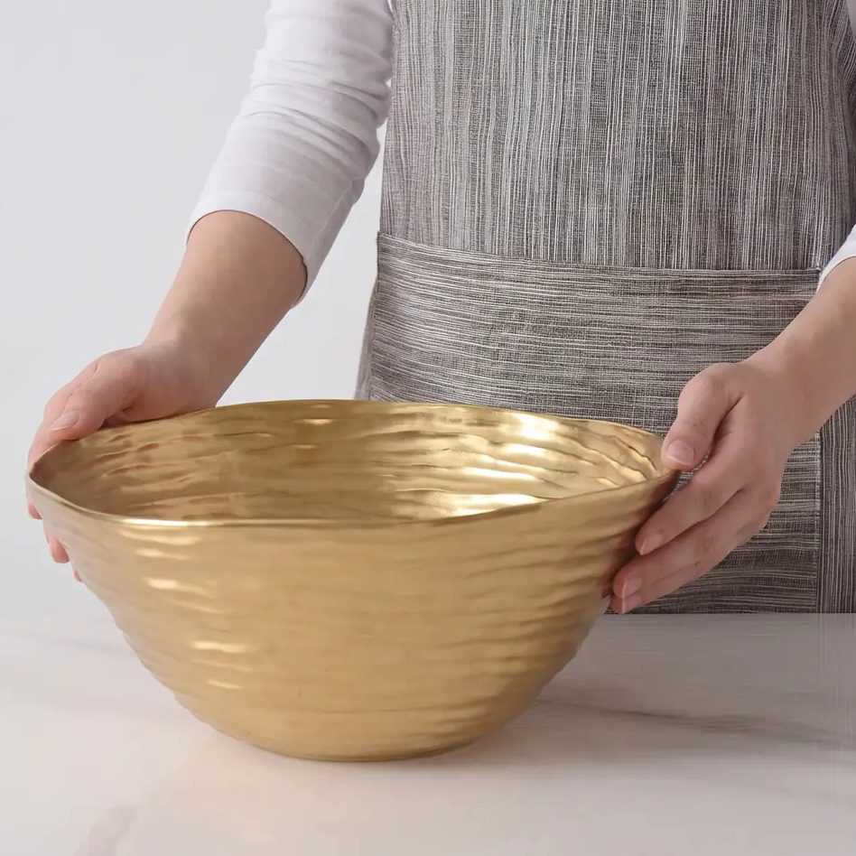 A person in a striped apron holds the Dorrato Gold Bowl - by PAMPA BAY with both hands over a white surface.