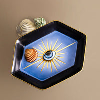 The L'Objet Lito Eye Hexagon Tray, a porcelain catchall featuring a blue and 24-karat gold sunburst and central eye motif, holds two seashells and rests on a beige surface with an additional shell beside it.