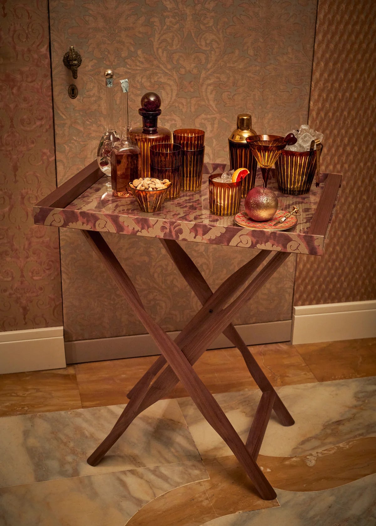 A wooden serving tray on foldable legs holds hand-crafted glassware, the L'Objet Prism Decanter, cocktail glasses, a bowl of nuts, and a teacup, set against a patterned wall and marble floor.