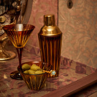 Amber hand-crafted glassware, a martini glass, the Prism Cocktail Shaker by L'Objet with a gold top, and a bowl of green olives rest on a decorative patterned surface.