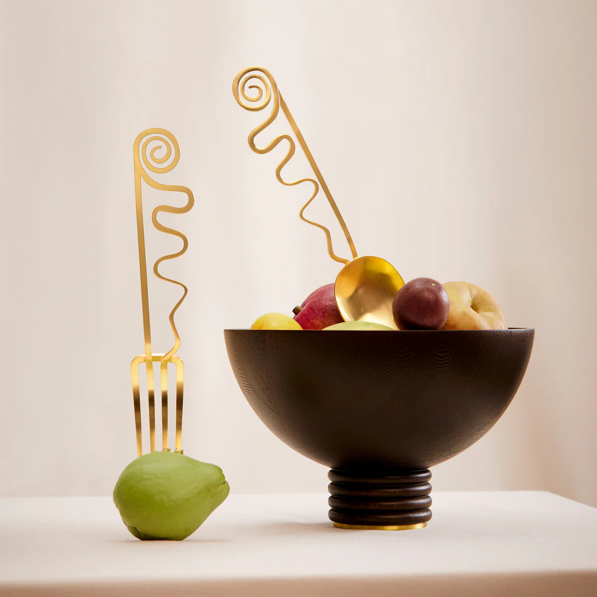 A modern still life showcases a black bowl of assorted fruit and a green pear, each accented with a large L'Objet Spirale Gold Serving Set utensil—a fork leaning on the pear and a spoon in the bowl—set against a beige background.