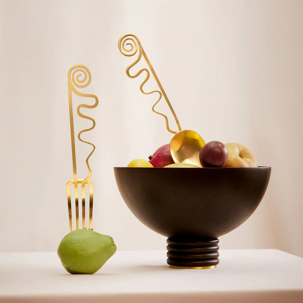 A modern still life showcases a black bowl of assorted fruit and a green pear, each accented with a large L'Objet Spirale Gold Serving Set utensil—a fork leaning on the pear and a spoon in the bowl—set against a beige background.