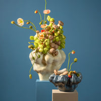 Two Timna Coupe vases by L’Objet, crafted in an aged iron finish and featuring wavy, organic forms, sit atop pastel blocks. Their stunning reactive glaze catches the light—the taller vase displays poppies and seed pods, while the smaller one holds just a scattering of pods. The scene is set against a solid blue background.