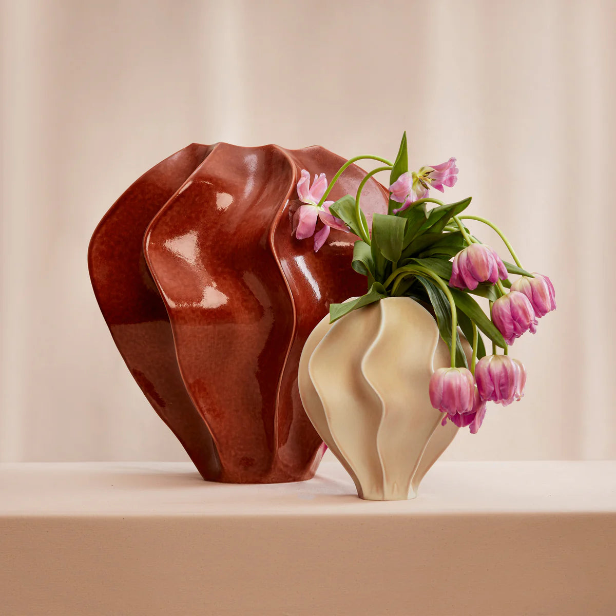 A glossy brown Douro Vase Paprika - Medium by L'Objet and a small ivory porcelain vase with pink tulips rest on a pale surface against a soft, neutral background—an elegant setting ideal for a luxury gift box display.