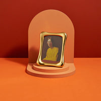 The Anton Frame Gold by L'Objet, featuring a wavy luxury design, displays a painting of a young woman in a yellow sweater with closed eyes. It is placed on a tiered pedestal before an orange arch.