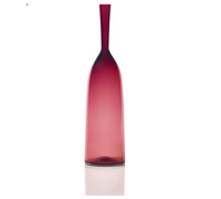 The Angelic Bottle - Small - by Cariati is a tall, slender red glass bottle with a narrow neck, shown upright on a reflective white surface against a plain background.