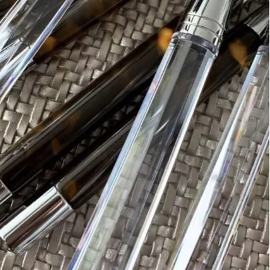 Several empty clear plastic lip gloss tubes with silver caps sit on a woven surface beside the Capdeco Doric Flatware 5 Piece Setting - Clear, creating an elegant and contemporary vibe in natural light.