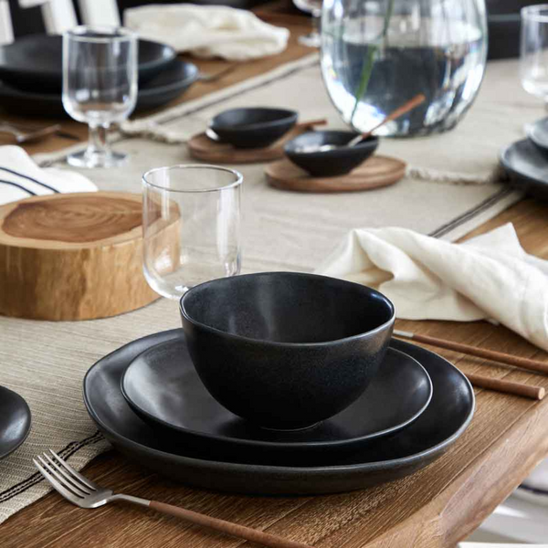 A modern dining setup with Casafina/Costa Nova Livia Dinnerware Matte Black, matching bowls, wooden-handled cutlery, clear glasses, cloth napkins, and decorative wood and glass accents on a beige table runner.