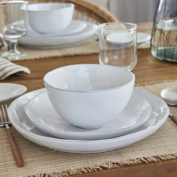 A close-up of a neatly set dining table featuring Casafina/Costa Nova’s Livia Dinnerware White, paired with glassware, cutlery, and a linen placemat on wood. Additional Livia place settings appear in the background.
