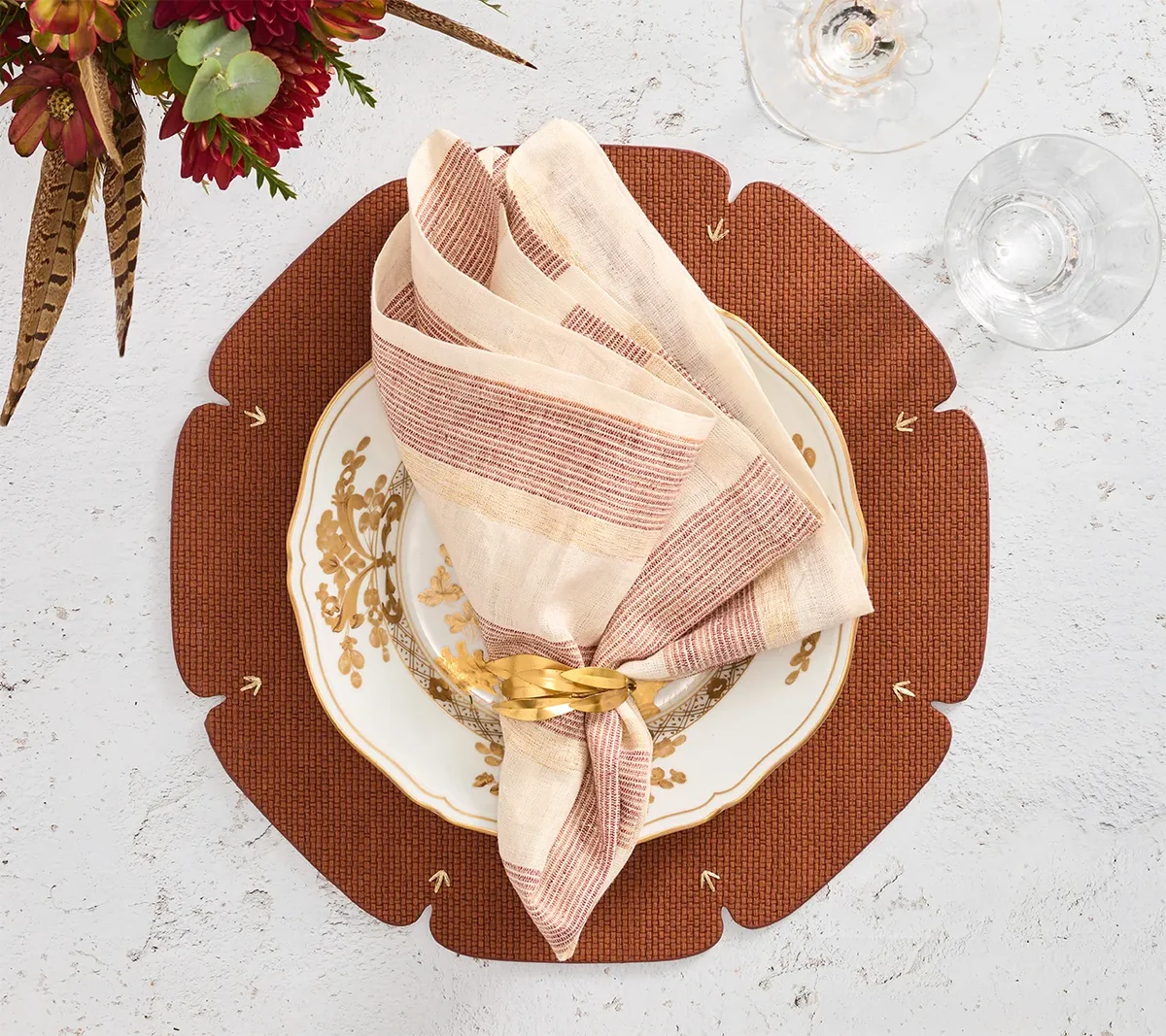 A decorative plate with gold patterns sits on a brown placemat. A brown and white Broad Stripe Napkin from Kim Seybert, held together with a gold napkin ring, is placed on the plate. Wine glasses and a floral arrangement are nearby on a white surface.