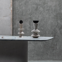Two Constantin Gray w/ Black Sculptures by Gardeco, featuring geometric forms and dark circular tops, sit on a marble-topped table against a gray textured concrete wall, embodying the essence of contemporary home decor and modernist sculpture.