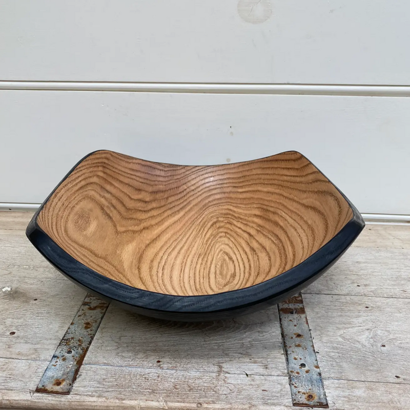 The Peterman Two Toned Square Oak Wood Bowl features smooth, curved sides and visible grain patterns. With a light brown interior, dark outer edge, and food-safe finish, it rests on a rustic wood surface against a white wall.