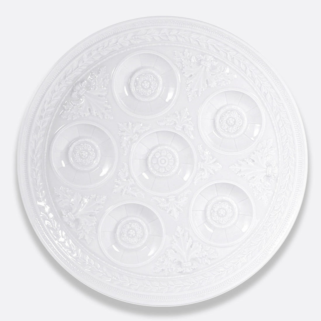 The Bernardaud Louvre Condiment Tray is a round, ornate white ceramic piece with seven circular indentations in a ring, featuring intricate decorative patterns inspired by French architecture—a refined Limoges dinnerware item.
