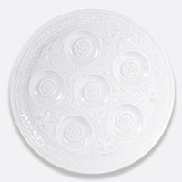 The Bernardaud Louvre Condiment Tray is a round, ornate white ceramic piece with seven circular indentations in a ring, featuring intricate decorative patterns inspired by French architecture—a refined Limoges dinnerware item.