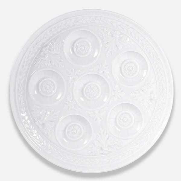 The Bernardaud Louvre Condiment Tray is a round, ornate white ceramic piece with seven circular indentations in a ring, featuring intricate decorative patterns inspired by French architecture—a refined Limoges dinnerware item.