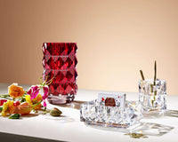 A red faceted vase, a Baccarat Louxor Catchall holding playing cards, a small crystal holder with gold pens, and scattered flowers sit on a white surface against a beige background.